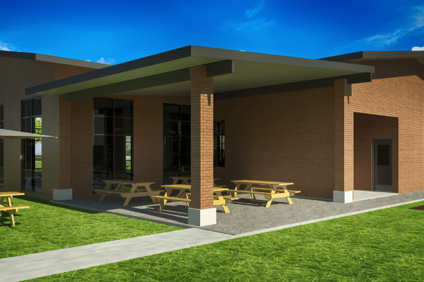 alt=’A covered patio with wooden picnic tables outside a brick building, set on a sunny day'