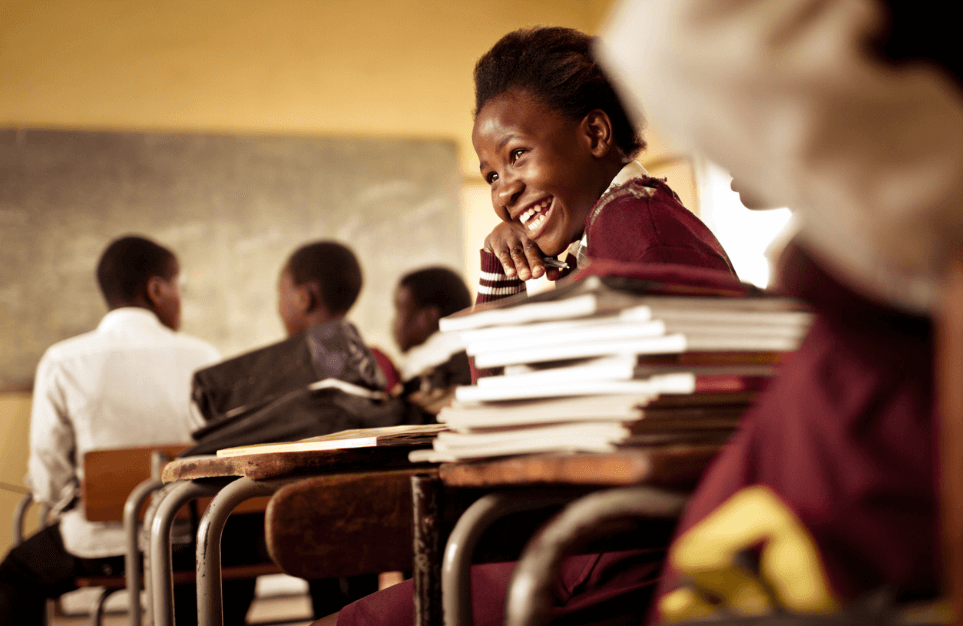 alt=’An African female student in uniform, beaming with joy while sitting at a desk with books, in a classroom setting'