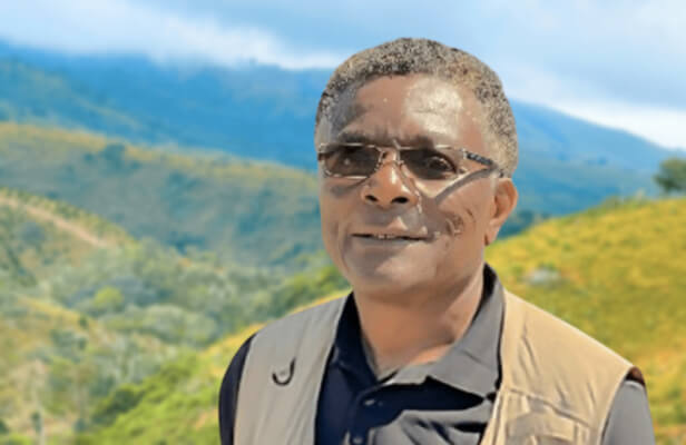 alt=’A man with glasses and a beige vest is photographed with scenic green hills and a cloudy sky behind him’