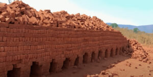 alt=’A brick kiln with neatly stacked red bricks and several archways, situated in an open field'