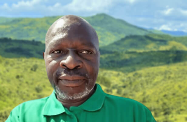 alt=’A man in a green shirt is photographed in front of a scenic landscape featuring hills and a cloudy sky'