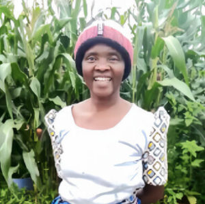 alt=’ A smiling woman in a white dress with patterned sleeves and a red hat stands amidst tall maize stalks'