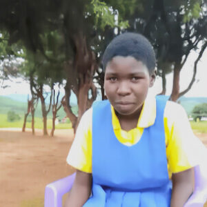 alt=’A young female student wearing a blue dress and yellow shirt, seated outside with trees and hills behind her'