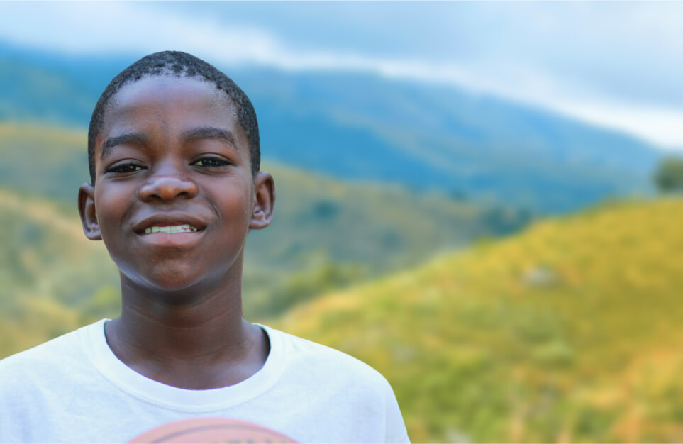 alt=’Portrait of a smiling boy with a backdrop of rolling green hills'