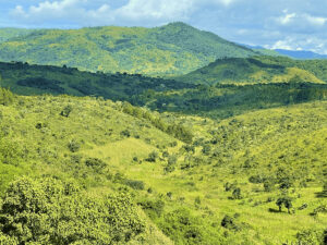 alt=’A panoramic view of lush green hills and valleys under a partly cloudy sky'