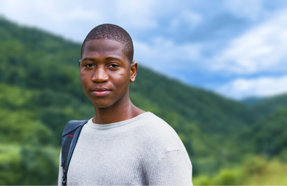 alt=’Portrait of a young man with a backpack, set against a scenic mountain backdrop'